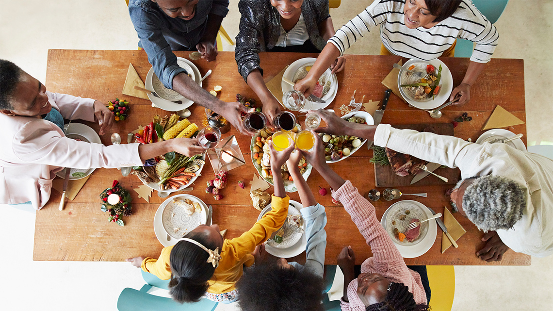 There are eight people sitting at a table that has plates and food on it. They are all reaching into the middle with their glasses.