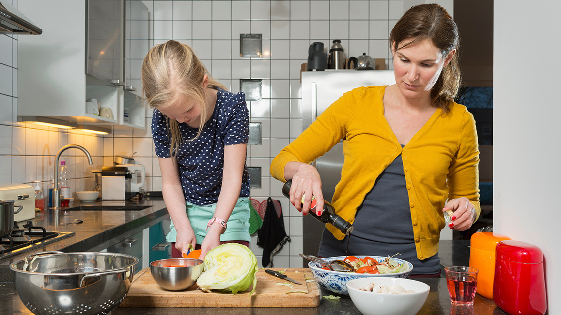 An adult and a child in the kitchen standing in front of a cutting board. There are vegetables in front of them and fruit.