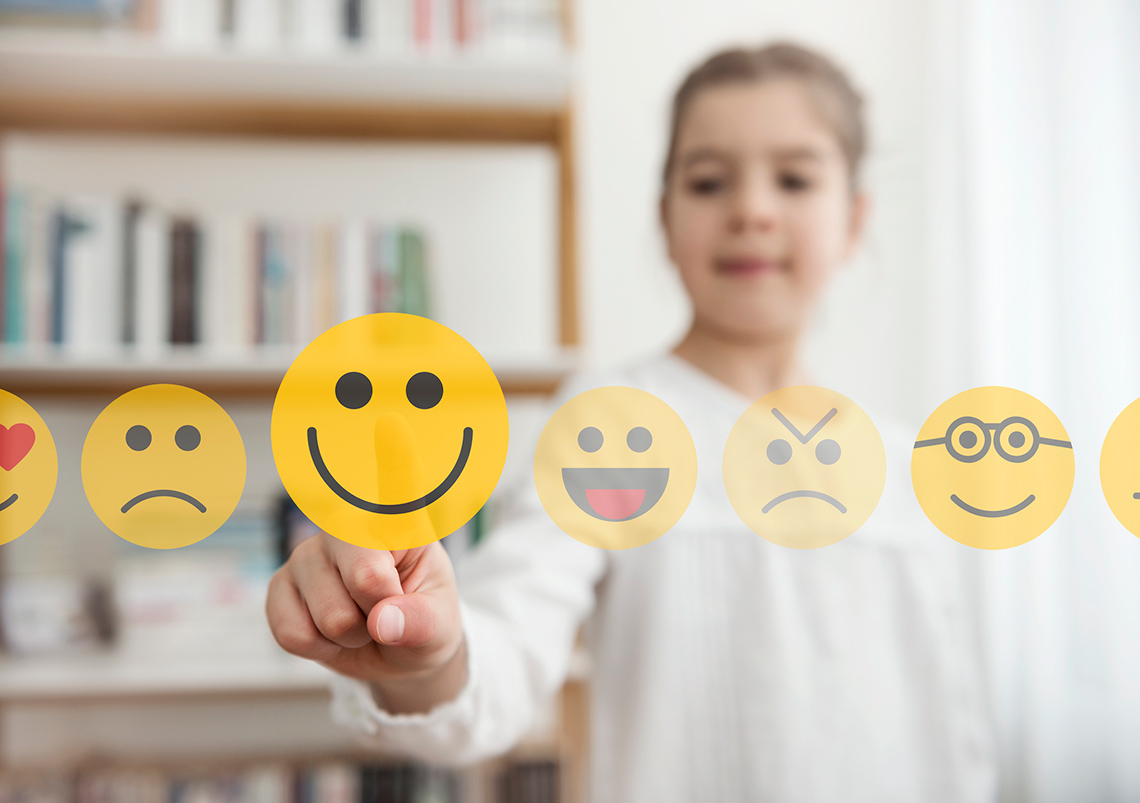 There is a child standing behind a line of faces with different emotions. The child is pointing to the smiling face.