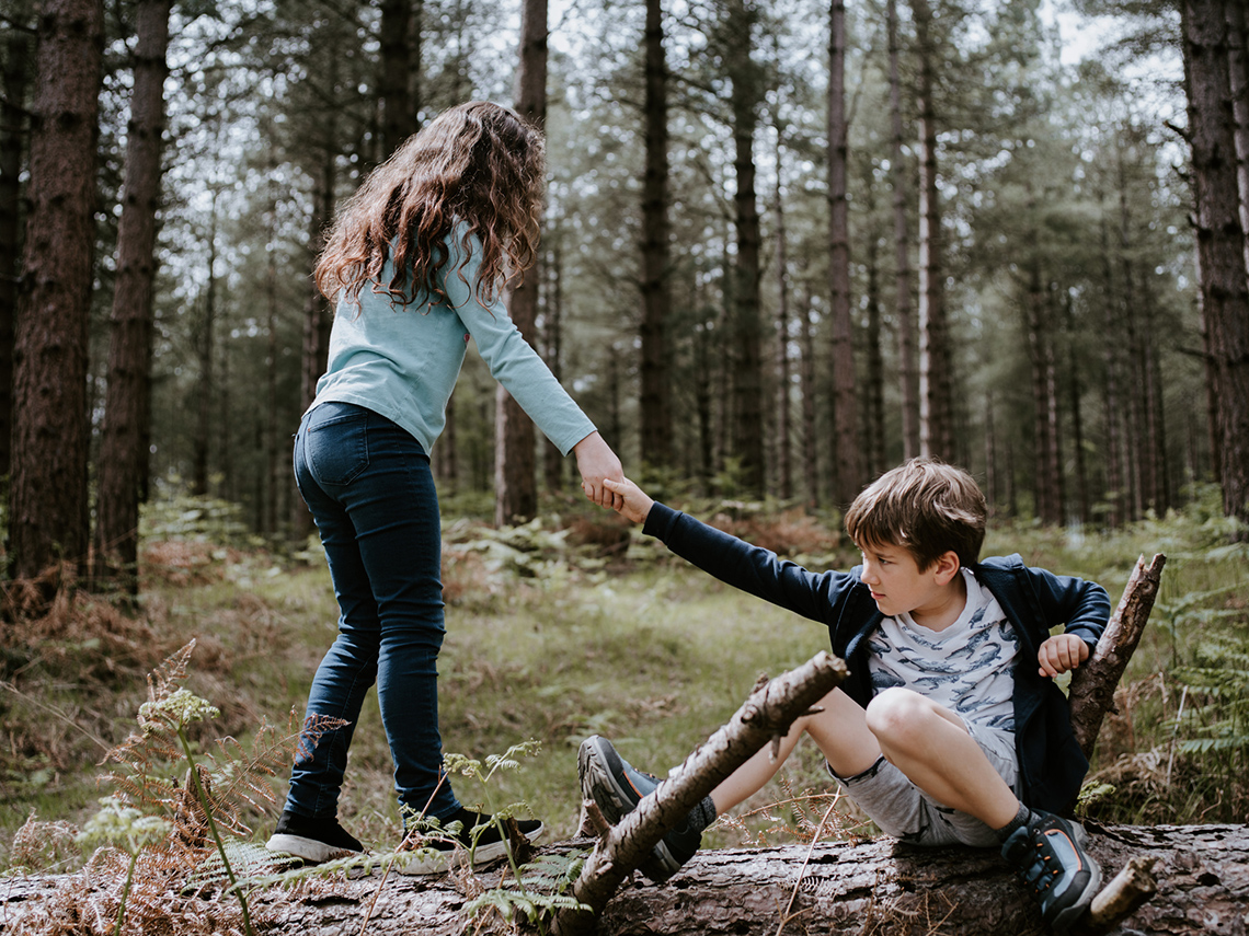 One child is on the ground while another child helps them up by holding their hand and pulling their arm.