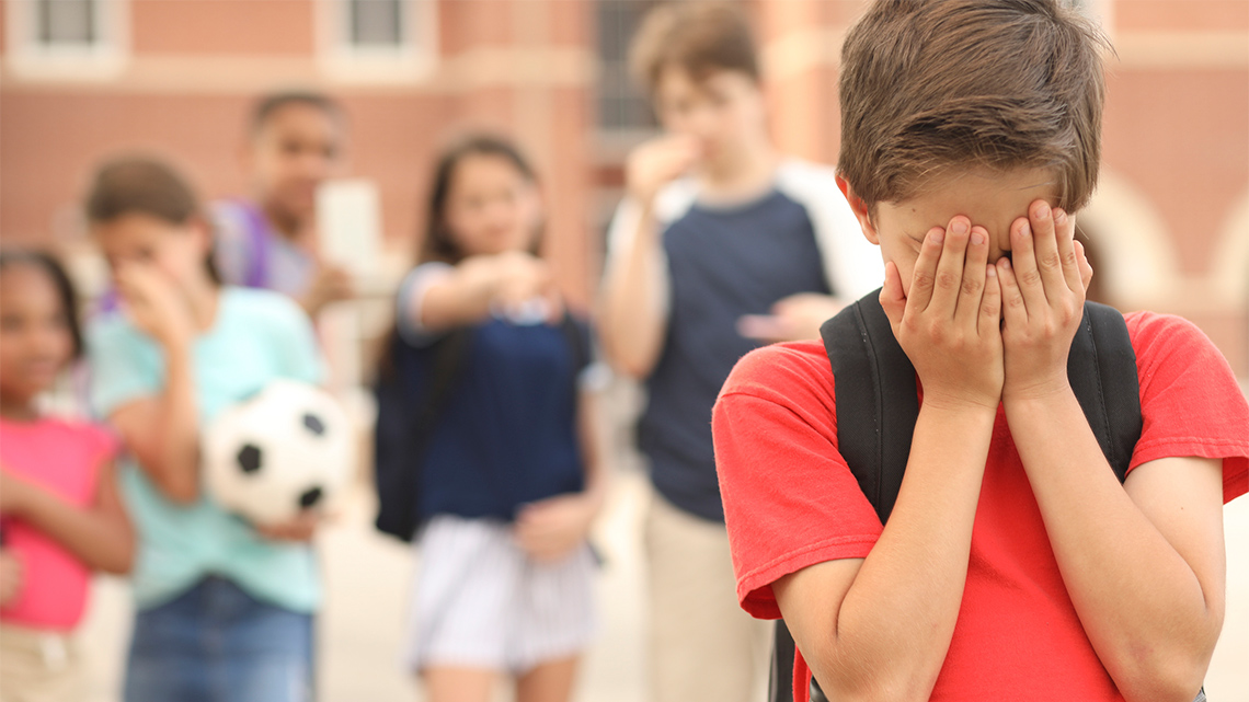 There are five kids in the background. One of the kids are pointing to the youth in the front who is covering their face with their hands.