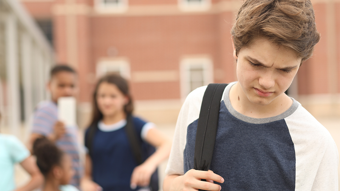 A youth looking down to the ground. A group of youths are behind them in the background filming on their phones.
