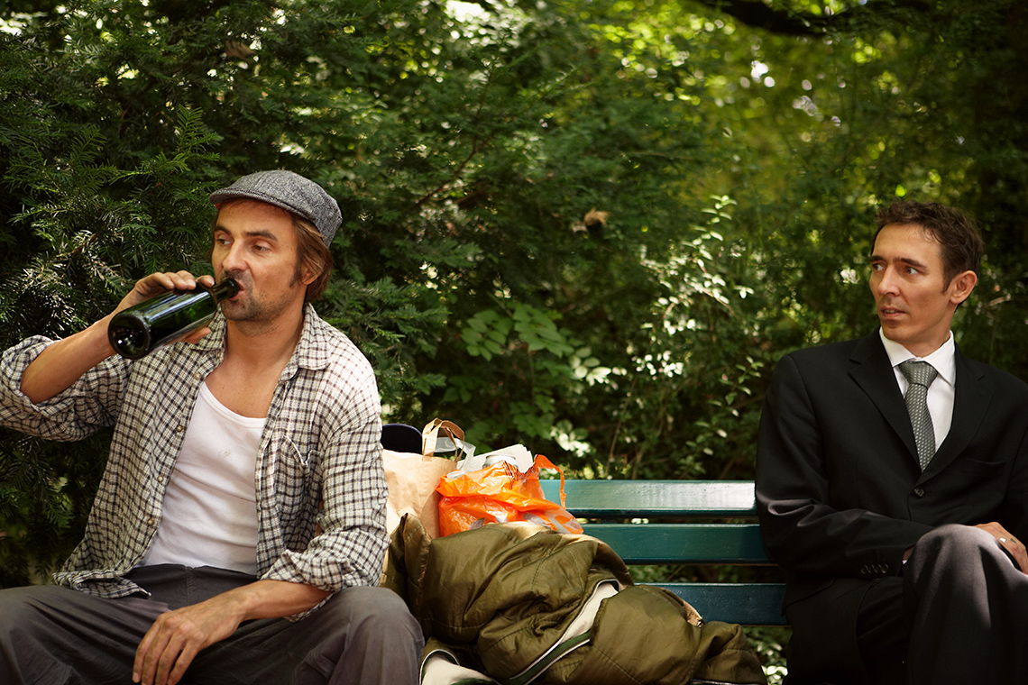 Two people sitting on a bench. One appears to be homeless and drinks a beer. The other is dressed in a suit, and stares at the other person.