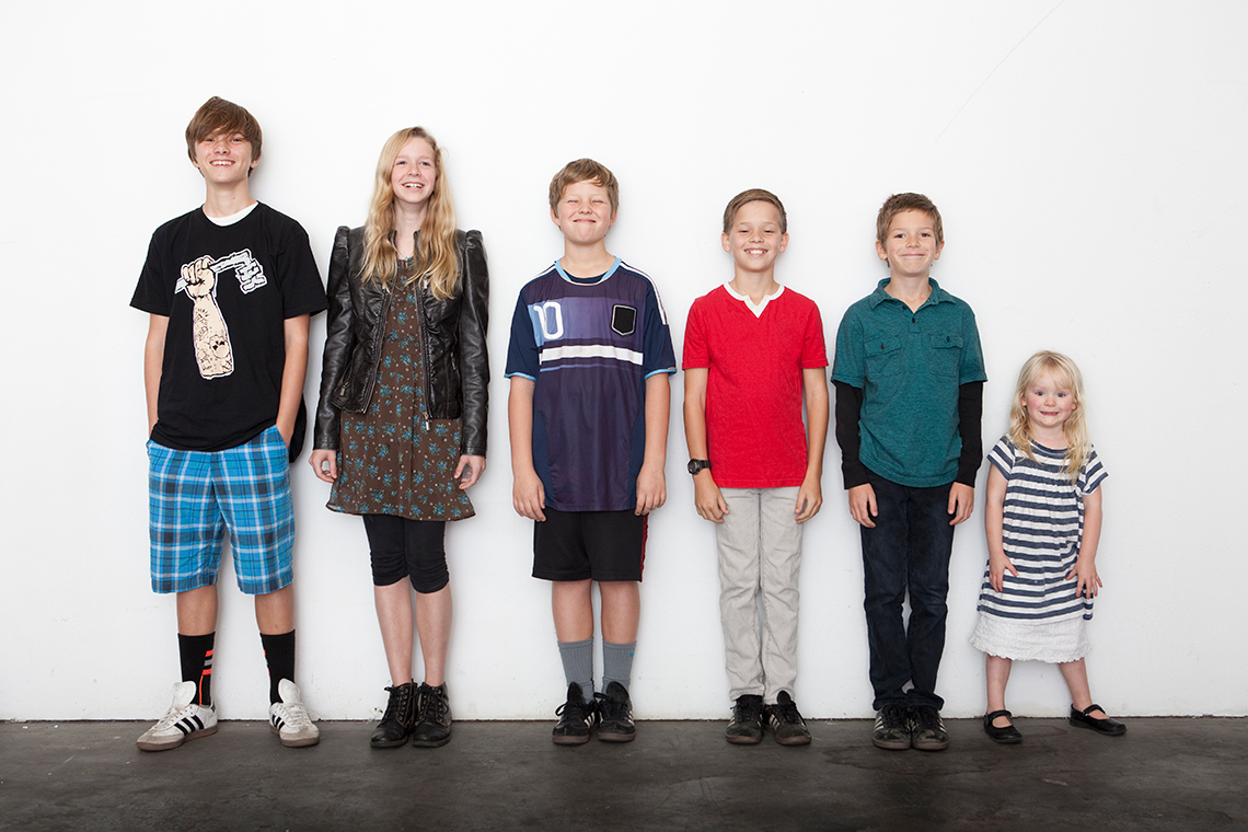 There are six children of different ages and heights standing against a wall.