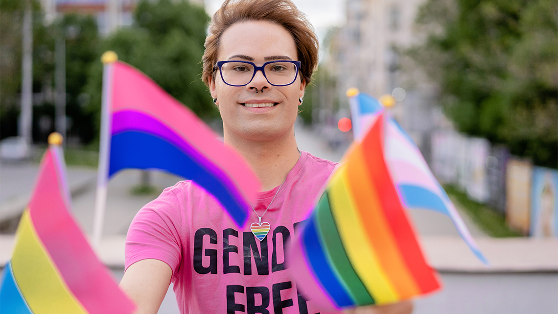 A non-gender binary individual holding a variety of pride flags.