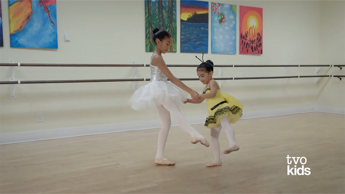 Two girls holding hands, and extending their legs to the side with their toes pointed.