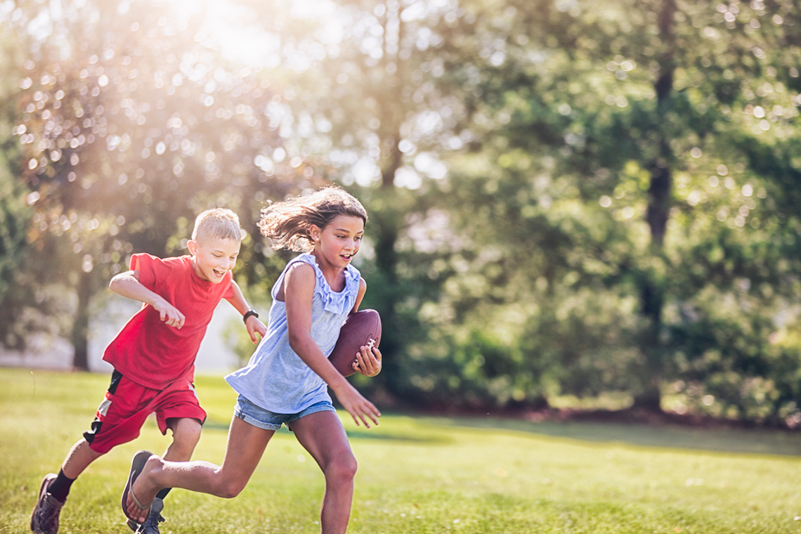 One child running and carrying a football while the other child follows behind.