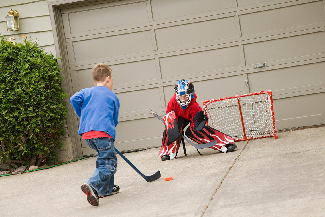Child hockey player keeping the puck close while moving towards the net on a driveway in front of a home. Another child is in goal.