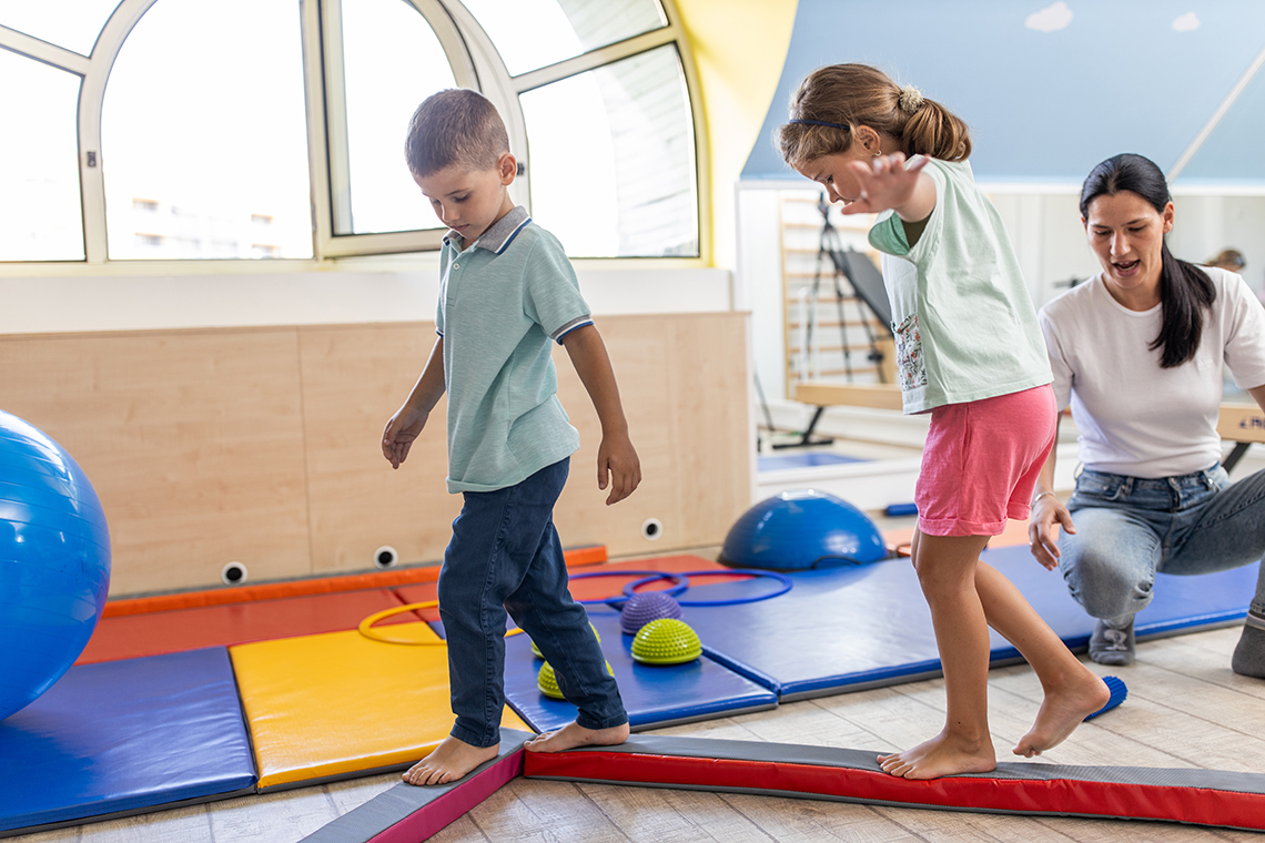 An adult instructs two children to walk along a low beam on the floor.
