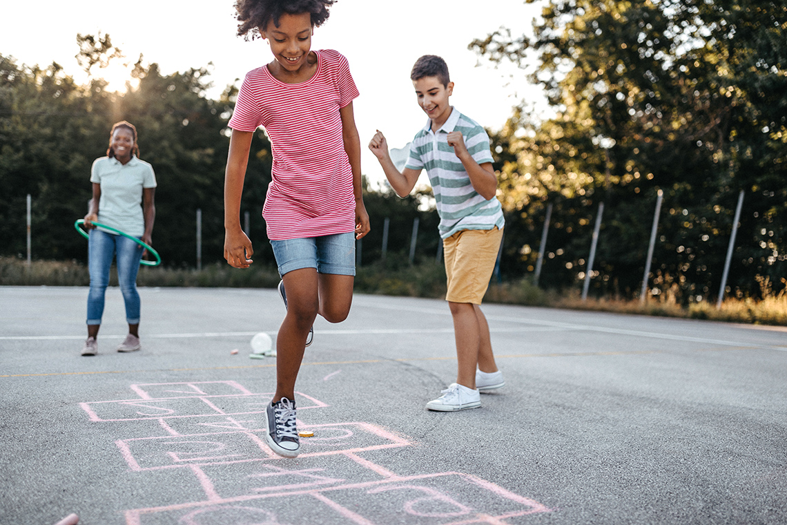 A child hopping on one foot playing hopscotch as their friends watch and cheer.