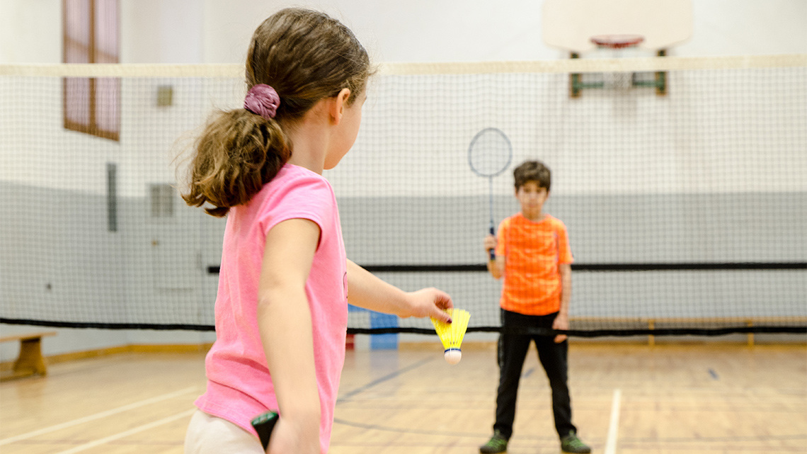 Children holding racquets on opposite sides of a net in a gym. One is swinging to serve a birdie.