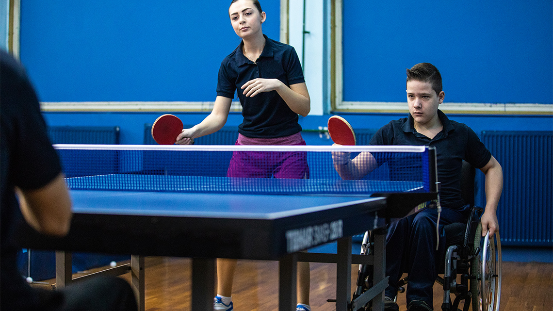 Two youth holding paddles on the same side of a small net on a table. One is using a wheelchair.