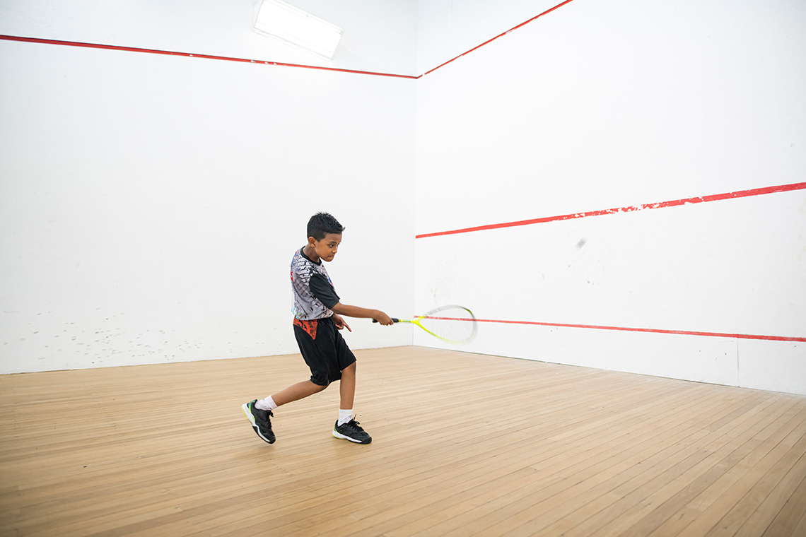 A child sending ball with a squash racquet towards a wall with red horizontal lines.