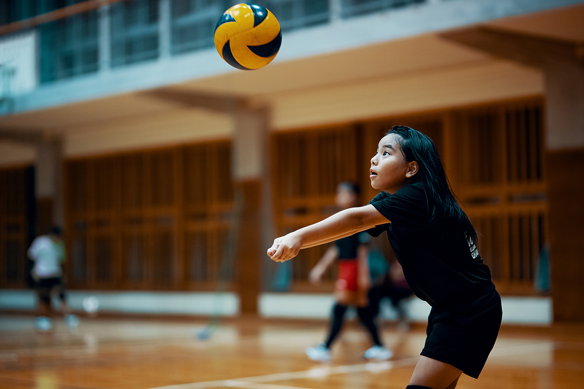 A volleyball falling toward a child clasping hands and stretching their arms toward the ball.