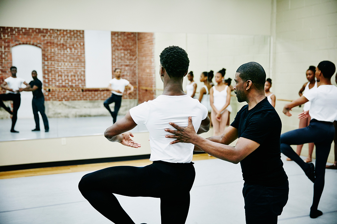 An instructor assisting a dancer to hold a pose on one foot.