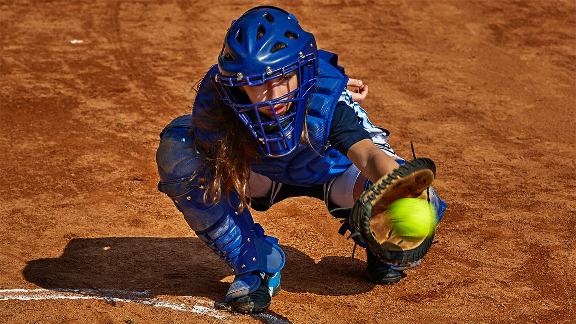 A person is crouched down preparing to catch a baseball. They hold a baseball glove outstretched in front of them.