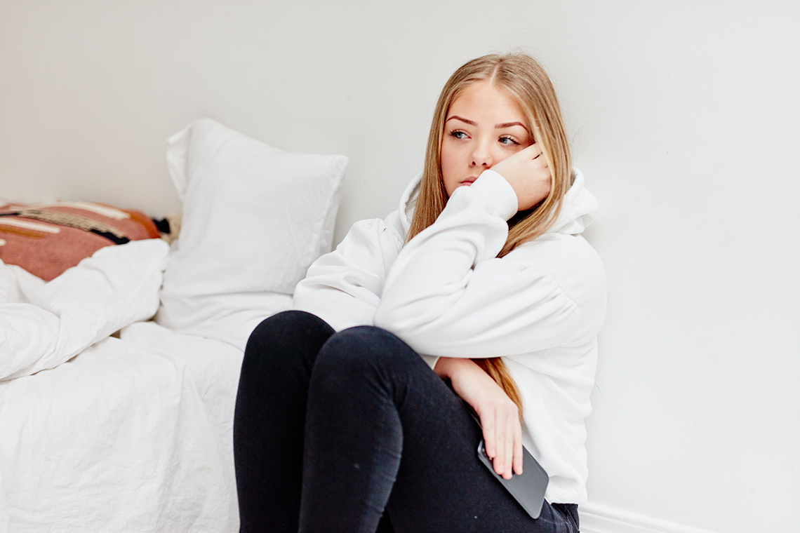 A girl looks sad. She sits on the floor with her knees tucked against her chest.