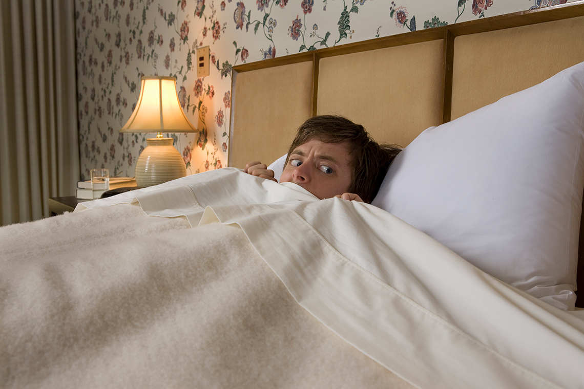 A person is in bed with the covers pulled up to their nose and a worried expression on their face.