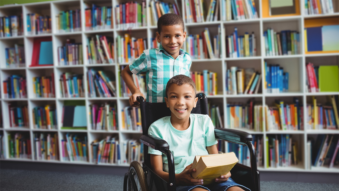 A child stands behind another child in a wheelchair, who is holding books in their lap.