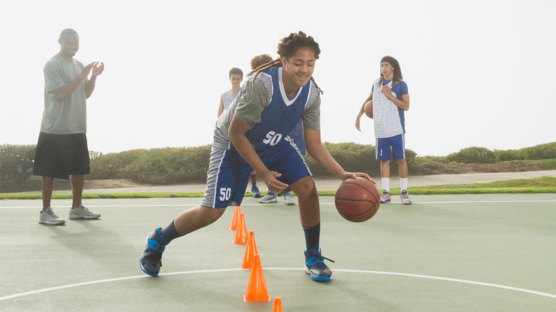 Several people dribble basketballs between their hands as they weave around practice cones.