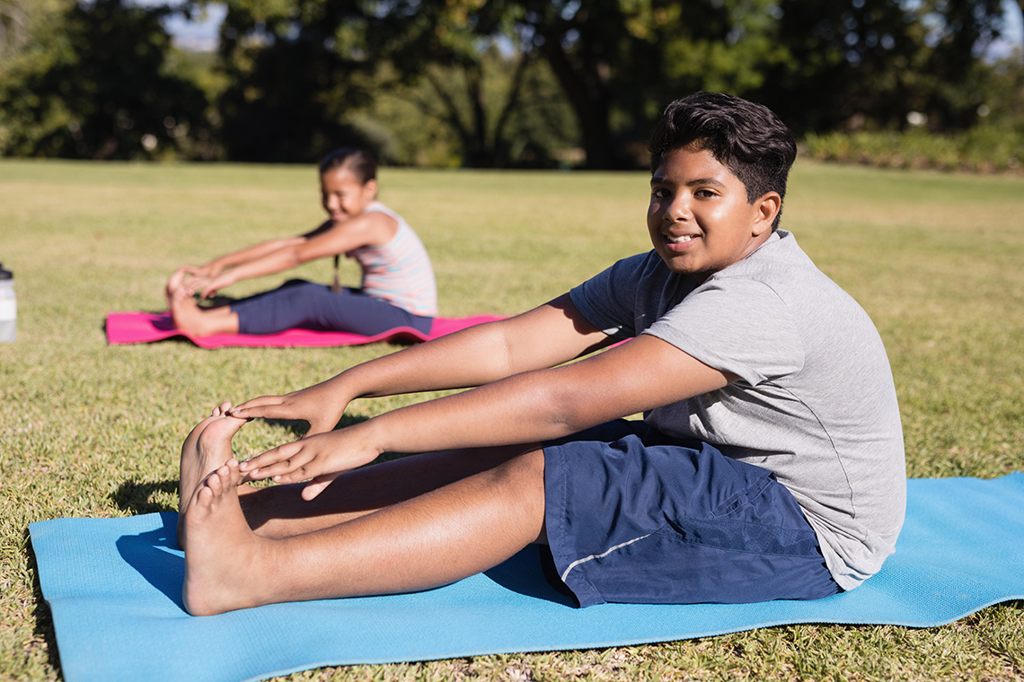 Two people stretch outside. They sit on the ground with their legs extended in front of them and reach towards their feet.