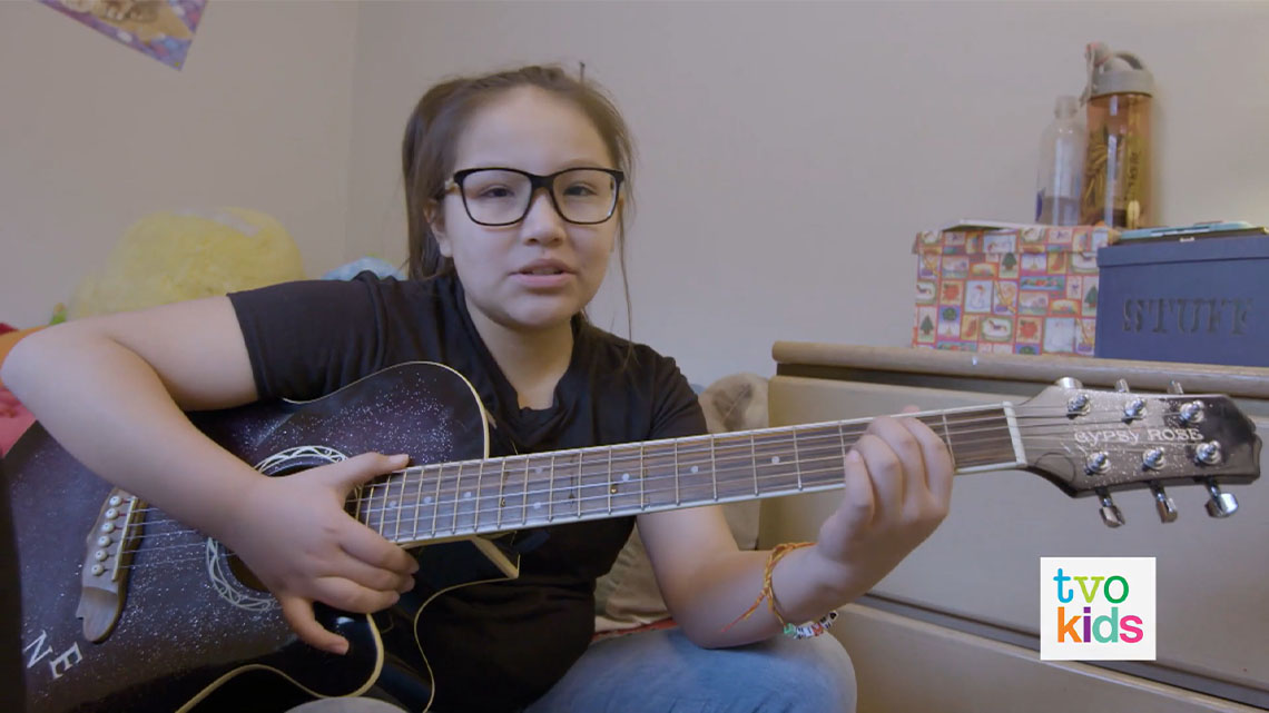 Adriana is playing a guitar in a room.