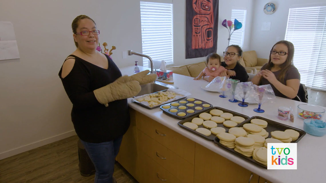 Natalya and Adriana are in a kitchen with their mom and baby sister. There are cookies and muffins on the counter.