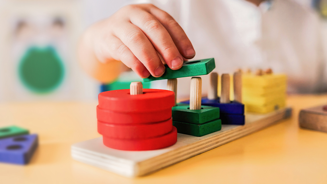 A child's hand puts a wooden shape on a peg toy.