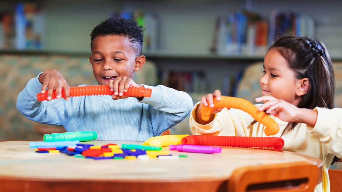 Two young children sit at a round table playing with bendable plastic tubes.