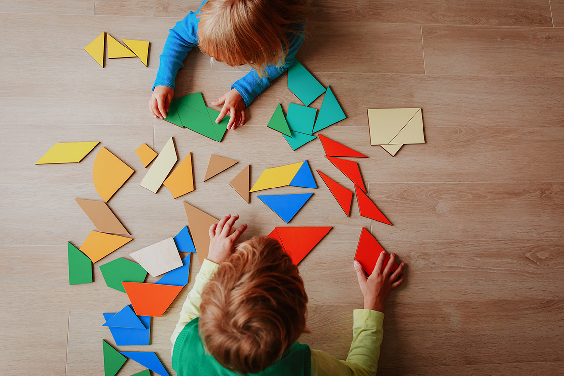 Two children sit on the floor arranging flat shapes.