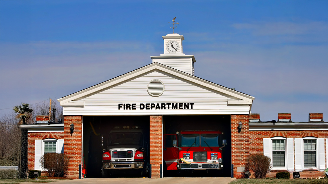A fire house is shown from the front. The building has flat walls, rows of windows, two open garage doors, and a clock tower on top.