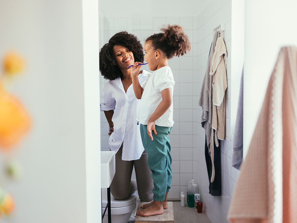 A young child stands on a stool brushing their teeth at a sink.