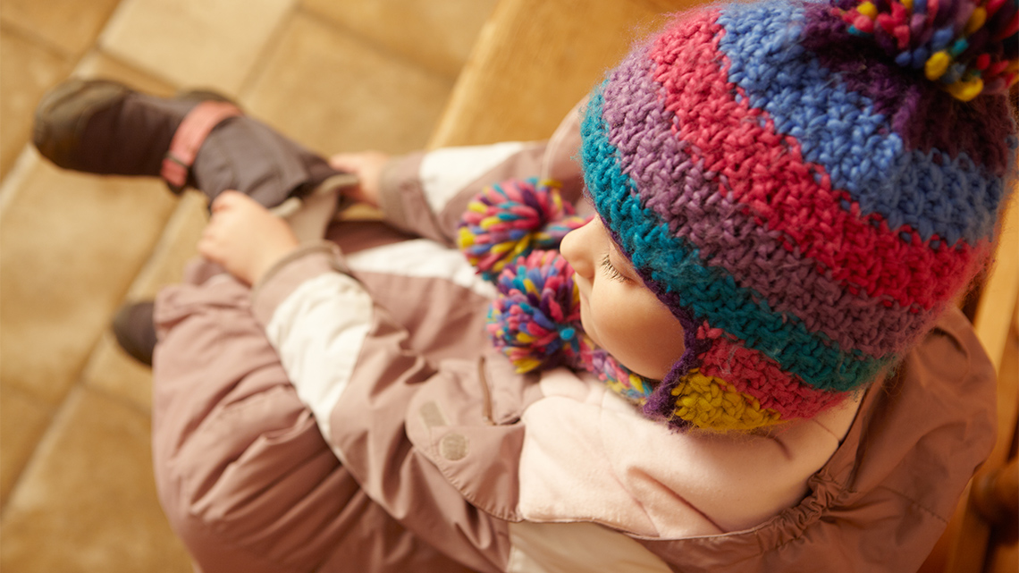 A child sits down and puts on a boot while wearing a jacket and a knit hat.