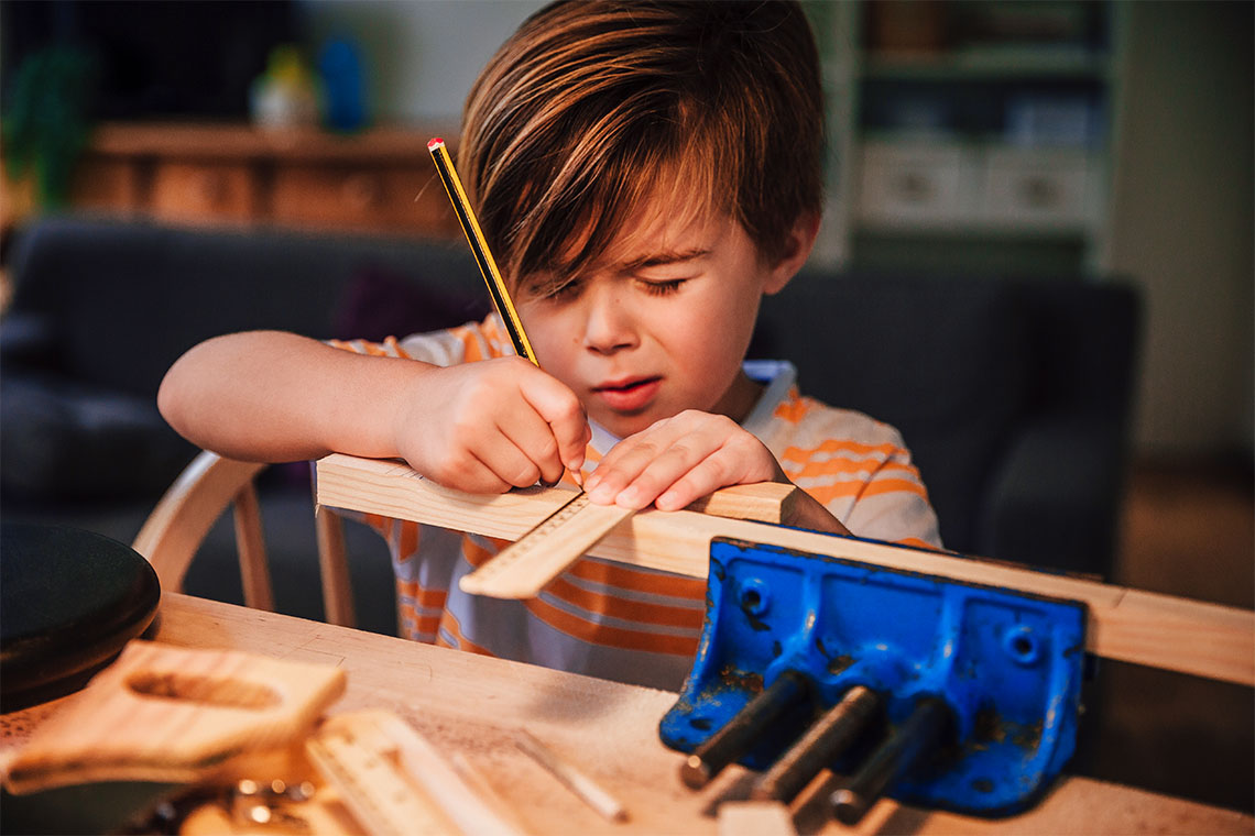 A child measures a piece of wood with a ruler while it is held in a clamp on a table.