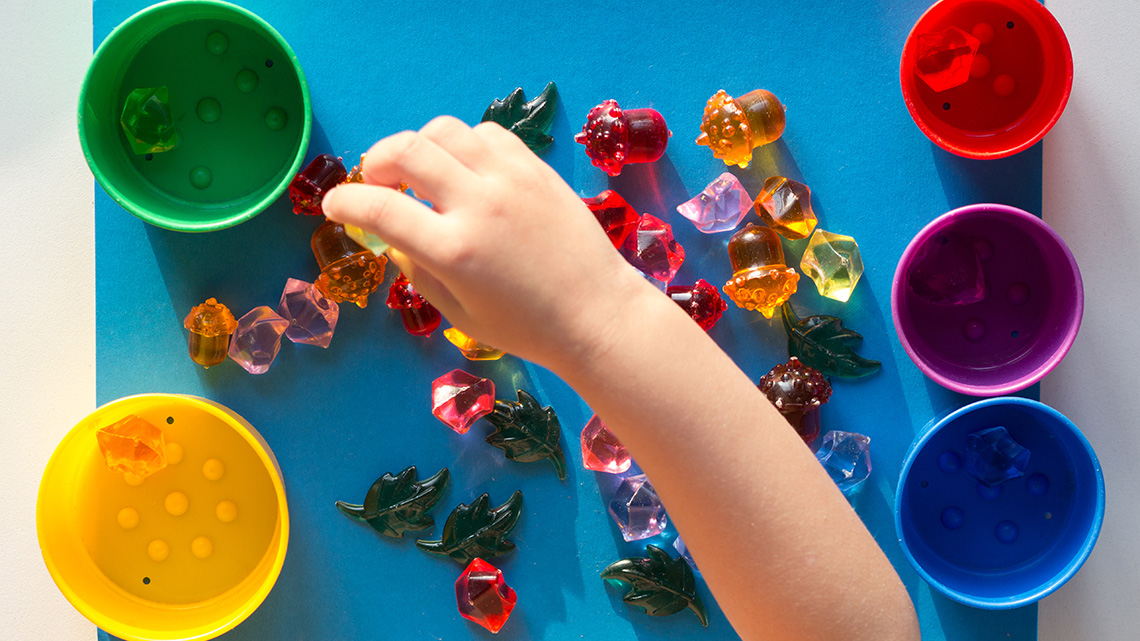 A child’s hand is separating multicoloured toy stones scattered on a table top into five separate cups.