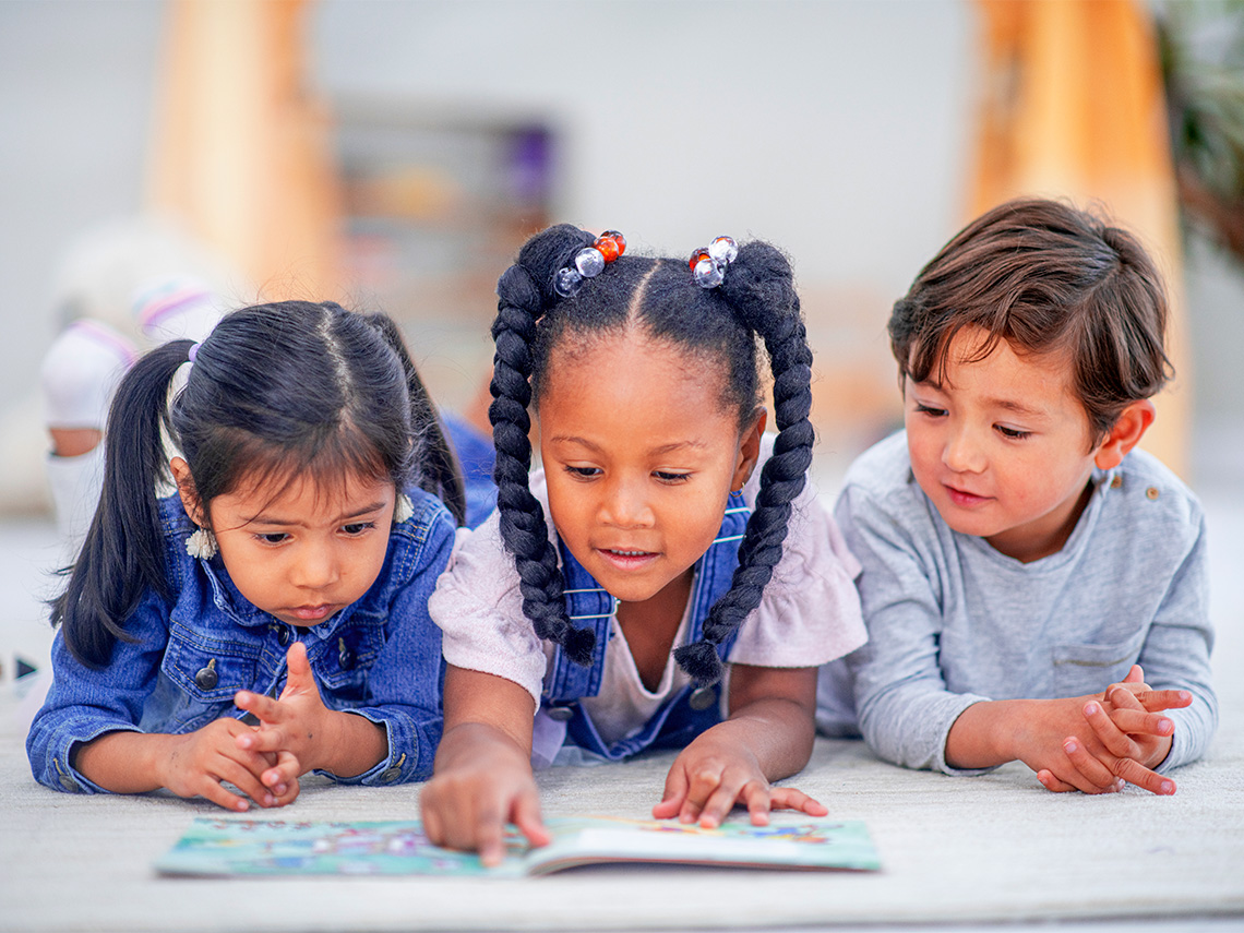Three young children laying on the ground reading a book together.