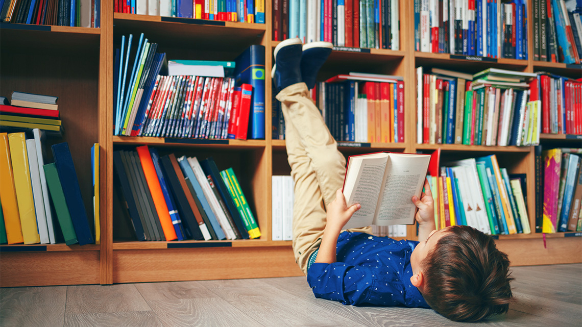 A child lies on the floor holding an open book, with their feet resting against a bookcase.