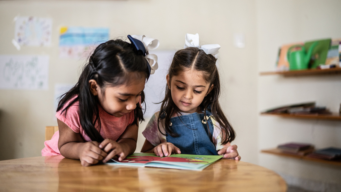 Two young children sit at a table while reading a book.