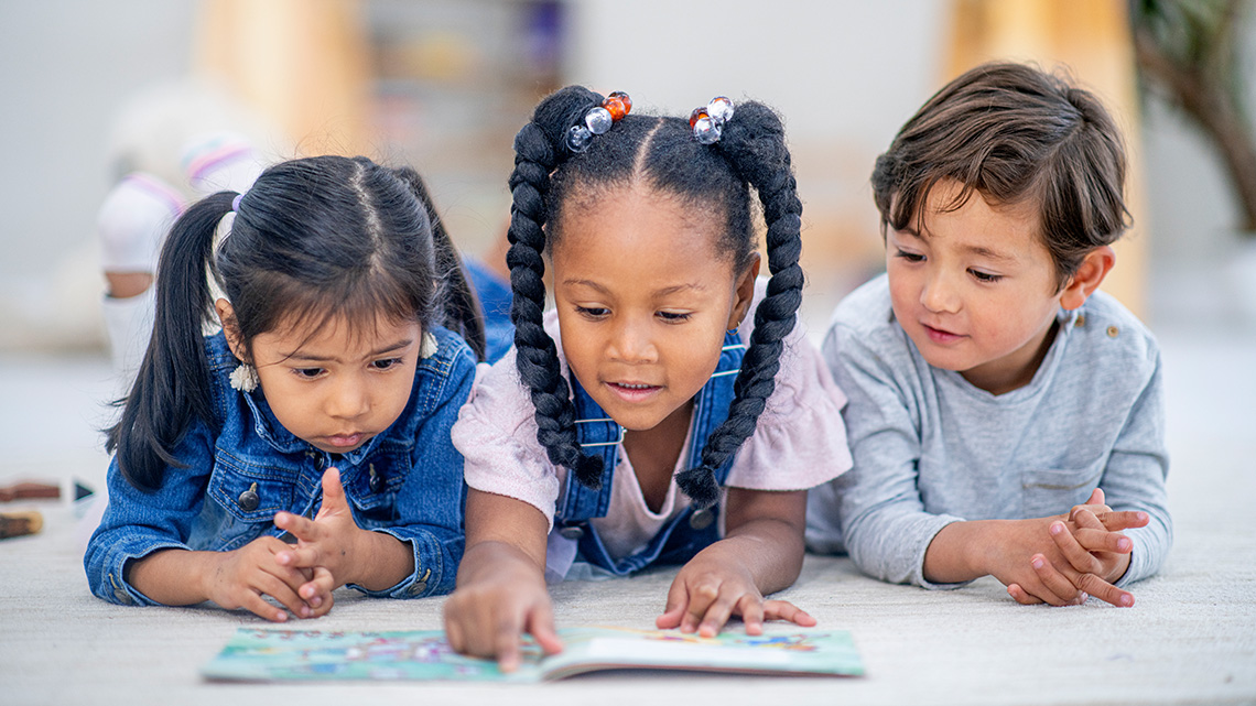 Three young children lie on the floor reading a book together.