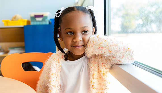 A young child rests their arm on the window ledge and faces the window.