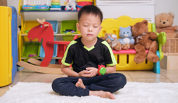 A young child sits with legs crossed and eyes closed on a rug.
