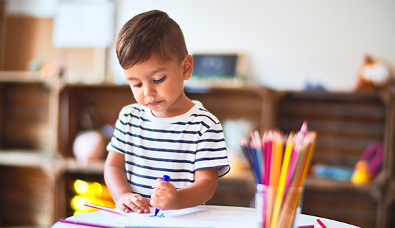 A young child draws on paper with a marker at a table.