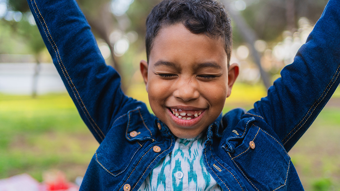 A young child is smiling with their eyes shut and arms raised above their head.