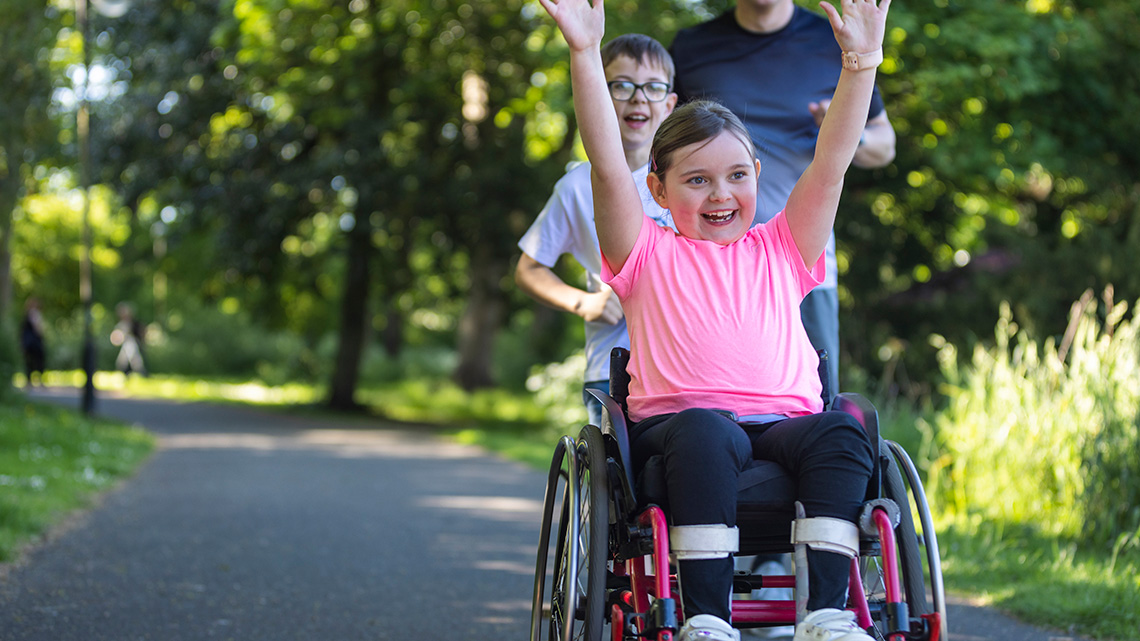 A young child in a wheelchair is smiling with their arms raised above their head.