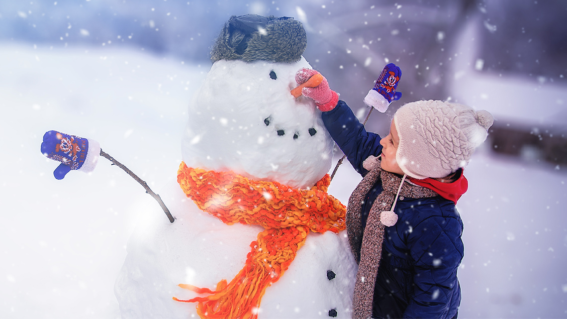 A child in a snowsuit builds a snowman outside.
