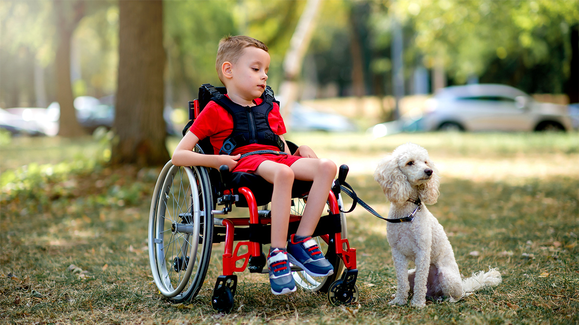 A little boy who uses a wheelchair sits in a natural space with his dog.