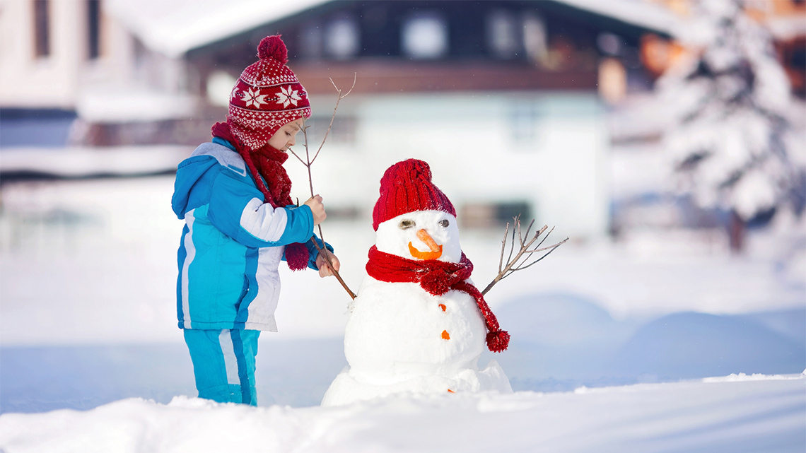 A child wears winter clothing and builds a snowman outside.