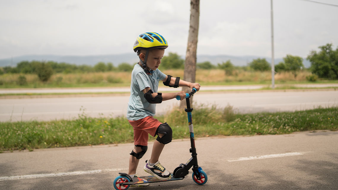Young child riding a scooter with a helmet, knee pads, and running shoes on