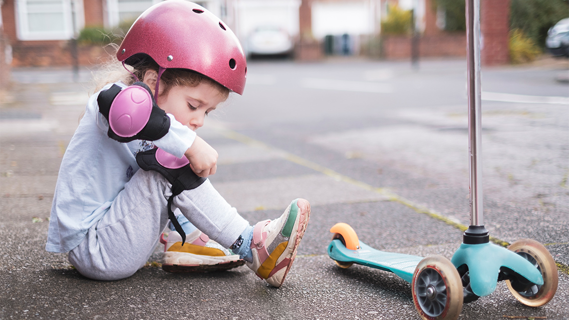 A child is getting ready to ride her scooter by putting on kneepads. They are wearing a helmet and elbow pads.