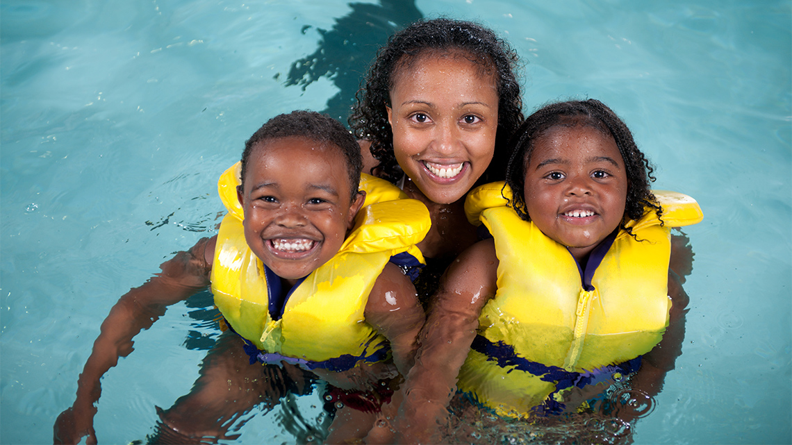 Two children in lifejackets are held by an adult in a swimming pool.